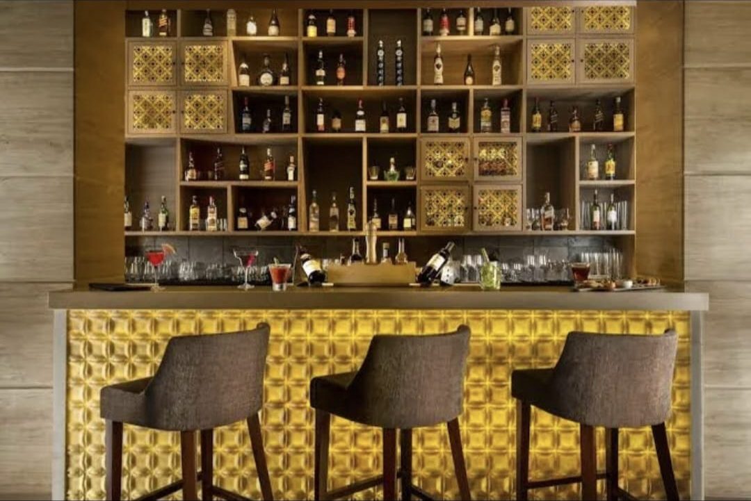Three upholstered bar stools at a backlit home bar counter with display shelving behind